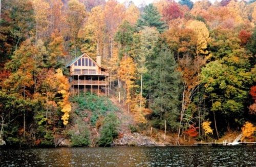 A cozy cabin on a hillside surrounded by vibrant autumn foliage and a calm lake.
