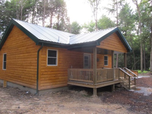 A wooden cabin with a metal roof, featuring a front porch and surrounded by trees.