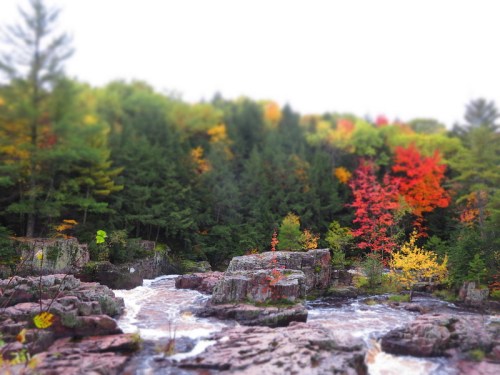 A serene river flows through rocky terrain, surrounded by vibrant autumn foliage in shades of red, orange, and yellow.