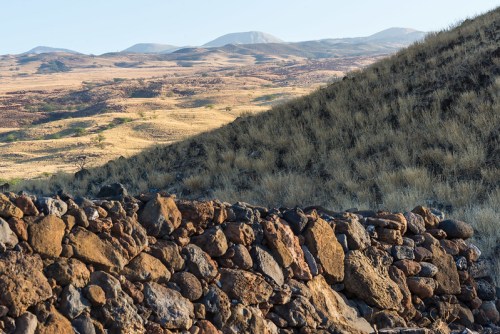 A rocky wall in the foreground with rolling hills and mountains in the background under a clear sky.