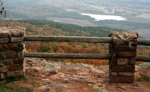 A scenic view from a rocky overlook, featuring autumn foliage and a distant lake surrounded by rolling hills.