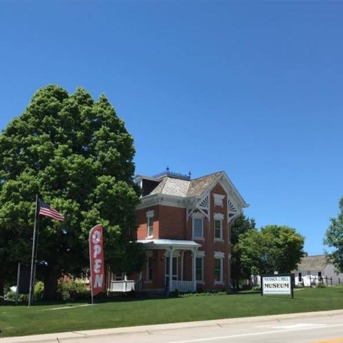 A historic brick museum building with a large tree, an "Open" flag, and an American flag under a clear blue sky.