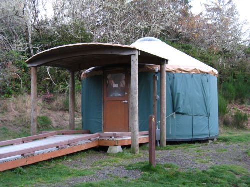 A green yurt with a wooden deck, surrounded by grass and trees, set in a natural outdoor environment.