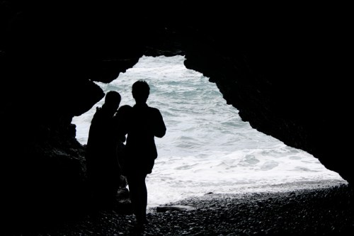 Silhouettes of two people standing in a cave, looking out at the ocean waves crashing on the shore.