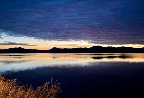 A serene lake at dusk, reflecting mountains and a colorful sky with clouds, creating a tranquil atmosphere.