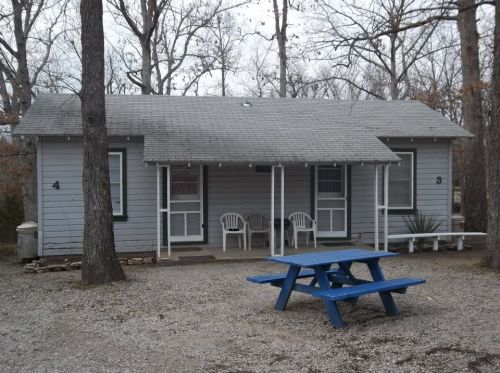A small gray cabin with a picnic table in front, surrounded by trees and gravel. Two doors and white chairs on the porch.