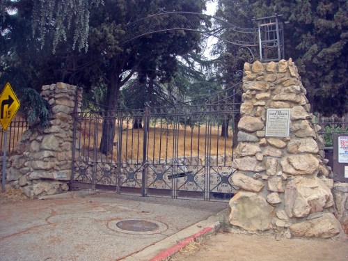Wrought iron gate with stone pillars, surrounded by trees and dry grass, leading to a park or private area.