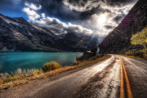 A winding road beside a turquoise lake, framed by mountains under a dramatic sky with sunlight breaking through clouds.