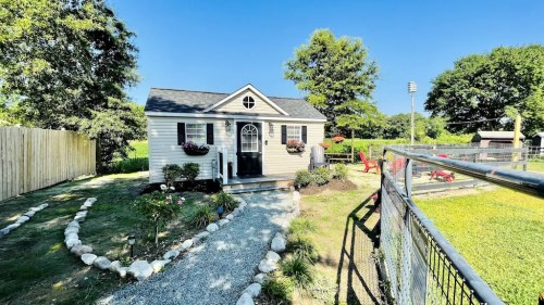 A small, charming house with a front porch, surrounded by a garden and a fenced yard on a sunny day.