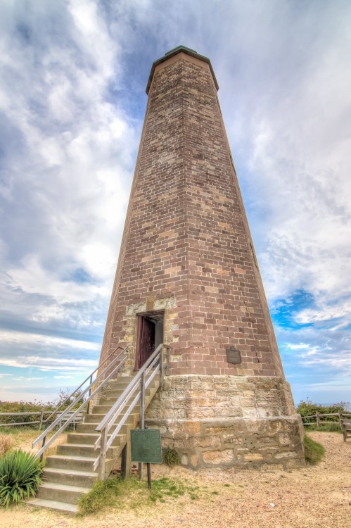 A tall stone lighthouse with a staircase leading to the entrance, set against a cloudy sky.