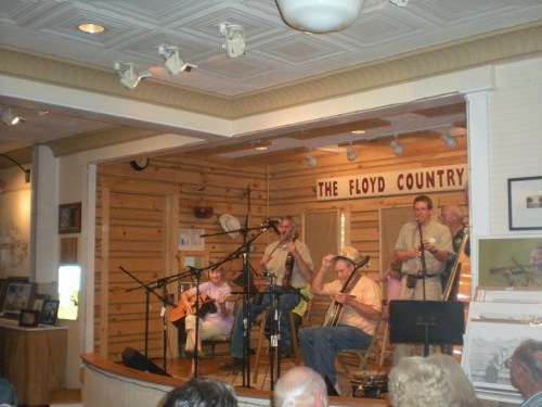 A band performs on stage in a cozy venue, with musicians playing guitar, banjo, and accordion. Audience in foreground.