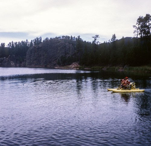 A yellow kayak with two people paddling on a calm lake surrounded by trees and rocky hills.