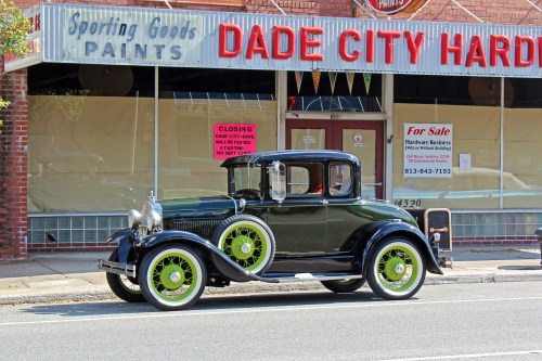 A vintage black car with green wheels parked in front of a hardware store in Dade City, Florida.