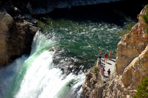 A scenic view of a waterfall with people standing on a viewing platform near the edge, surrounded by rocky cliffs.