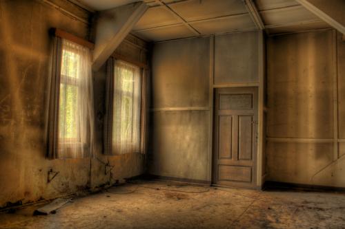 An empty, dimly lit room with two windows and a closed door, featuring worn walls and a dusty floor.