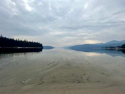 A calm lake reflects a cloudy sky, surrounded by trees and mountains, with gentle ripples on the sandy shore.