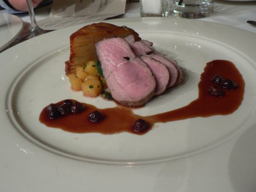 A plate featuring sliced roast meat, accompanied by diced vegetables, a crispy potato, and a berry sauce drizzle.