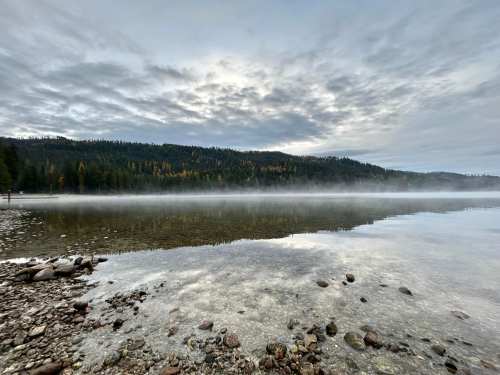 A serene lake scene with mist rising, surrounded by trees and a cloudy sky, reflecting the landscape on the water's surface.