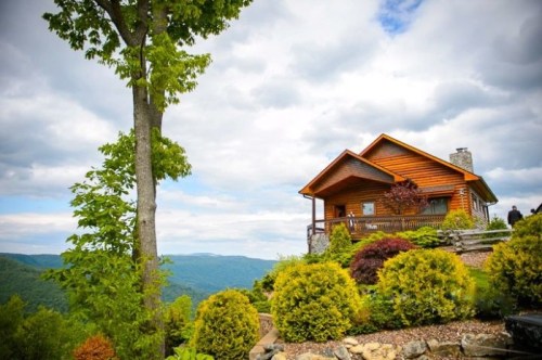 A wooden cabin on a hillside, surrounded by lush greenery and mountains under a cloudy sky.