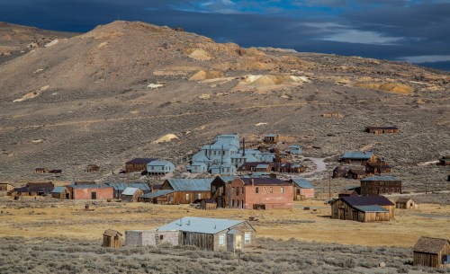 A ghost town with wooden buildings set against a mountainous landscape under a cloudy sky.