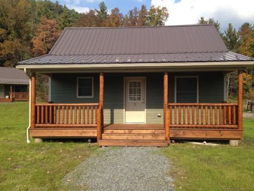 A small, green cabin with a metal roof, wooden porch, and surrounded by trees and grass.
