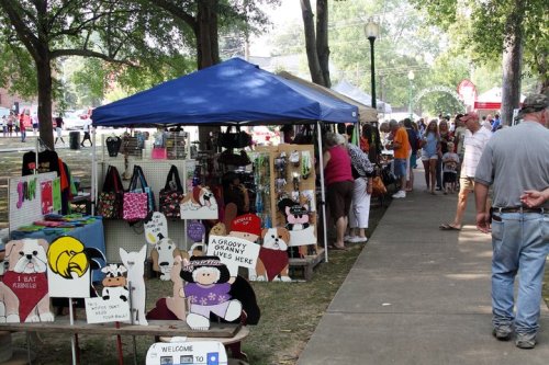 A bustling outdoor market with colorful stalls selling crafts and decorations, surrounded by trees and visitors.