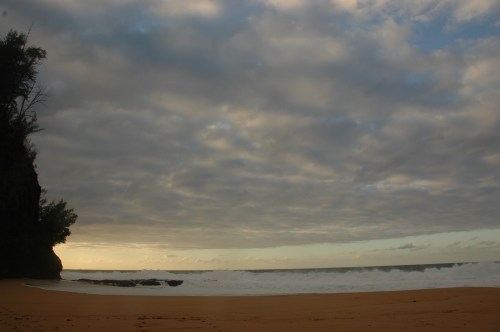 A serene beach scene with gentle waves, a rocky cliff, and a cloudy sky at sunset.