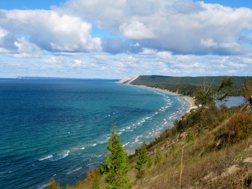 A scenic view of a coastline with blue waters, sandy shores, and green hills under a partly cloudy sky.