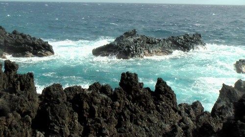Waves crash against rocky formations in a vibrant blue ocean under a clear sky.