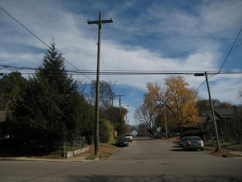 A quiet street scene with power lines, trees, and cars under a partly cloudy sky.