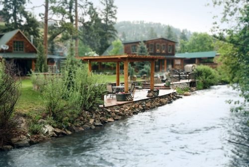 A serene riverside scene featuring a wooden deck with seating, surrounded by lush greenery and rustic buildings in the background.