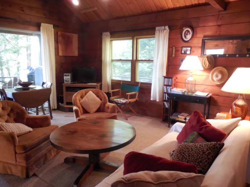 Cozy living room with wooden walls, a round table, two armchairs, a sofa, and large windows overlooking greenery.