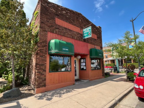 A brick building with green awnings and a sign, surrounded by trees and parked cars on a sunny day.