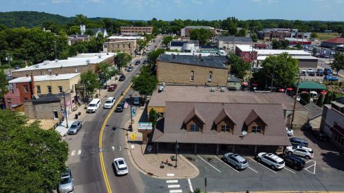 Aerial view of a small town with shops, trees, and parked cars along a main street on a sunny day.