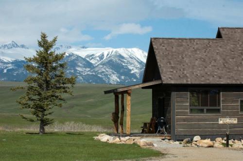 A rustic cabin with a porch, set against a backdrop of snow-capped mountains and green fields under a blue sky.