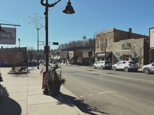 A sunny street view of a small town with shops, parked cars, and a water tower in the background.
