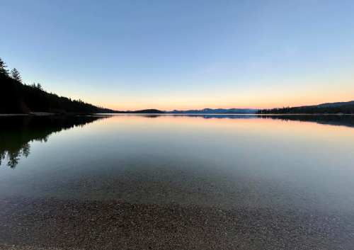 Calm lake at dusk, reflecting trees and mountains under a soft gradient sky. Peaceful and serene atmosphere.
