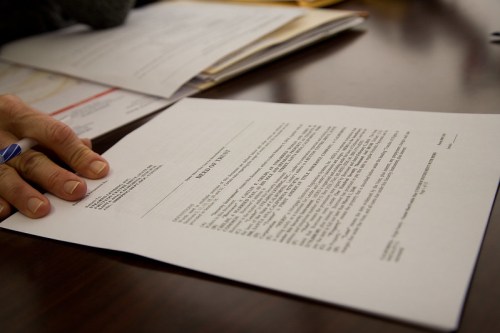 A hand rests on a document with text, placed on a wooden table surrounded by other papers.
