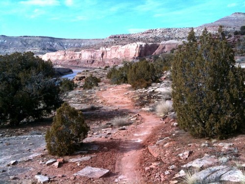 A winding dirt path leads through rocky terrain and sparse vegetation, with cliffs in the background under a blue sky.