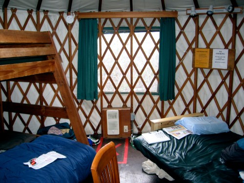 Interior of a yurt with wooden furniture, bunk beds, and a window covered by green curtains.
