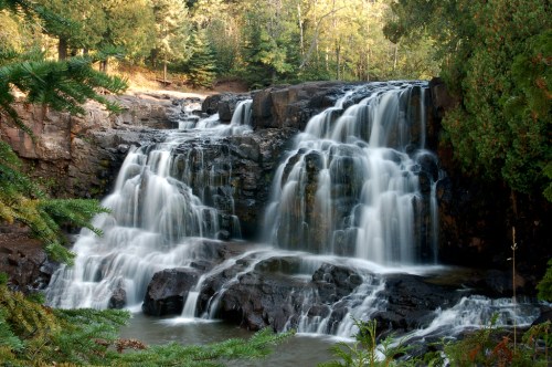 A serene waterfall cascading over rocky ledges, surrounded by lush green trees and a tranquil pool below.