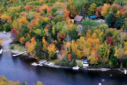 Aerial view of a colorful autumn landscape with trees in vibrant hues, a calm lake, and a few houses along the shore.