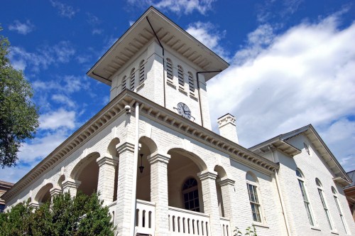 A white brick building with a clock tower, featuring arched windows and a blue sky with clouds in the background.