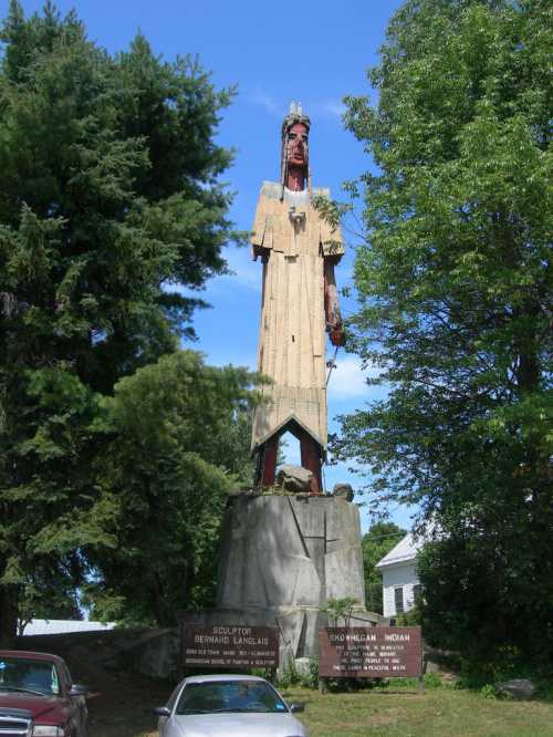 A tall wooden statue of a Native American figure stands on a stone base, surrounded by trees and parked cars.