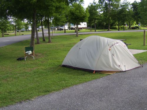 A tent set up on grass near a camping area, with a folding chair nearby and trees in the background.