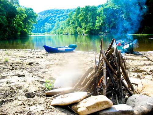 A campfire made of sticks and stones by a riverbank, with a kayak nearby and lush green trees in the background.