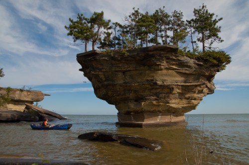 A small boat near a unique rock formation topped with trees, surrounded by calm water and a blue sky.
