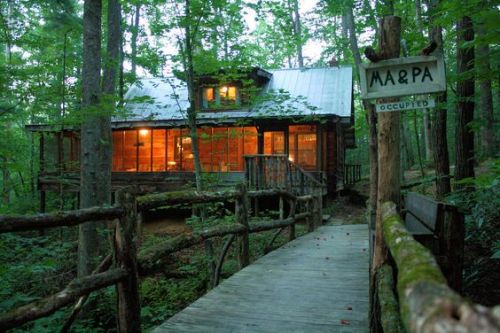 A rustic cabin in a forest, with large windows glowing warmly, connected by a wooden walkway and surrounded by trees.