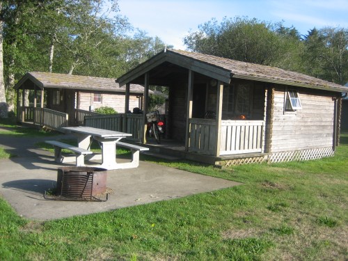 Two wooden cabins with a picnic table and fire pit in a grassy area, surrounded by trees.