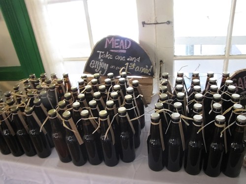 A table with numerous bottles of mead, each tied with string, next to a sign inviting guests to take one.